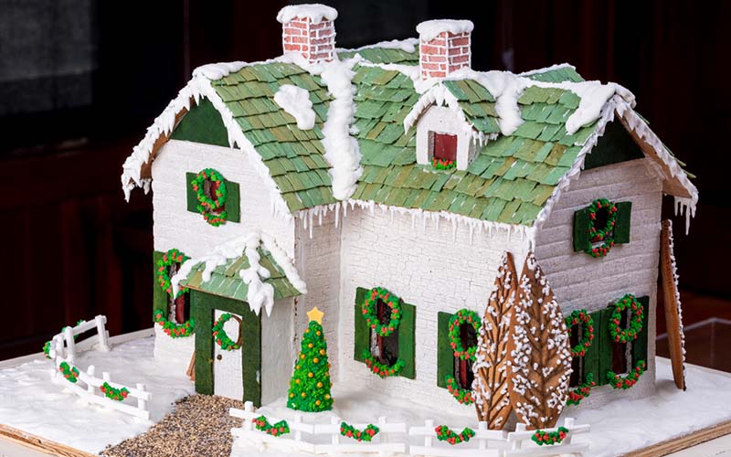 Photograph of a house covered with snow made from gingerbread and other baked goods. The house is white with a green roof. Fresh fallen snow covers part of the roof and ground. The house is inspired by Anne of Green Gables. 